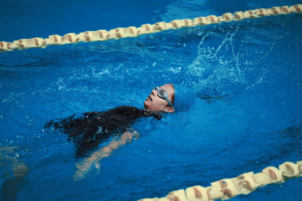 A young woman swims backstroke in an Olympic-sized swimming pool under clear blue water.